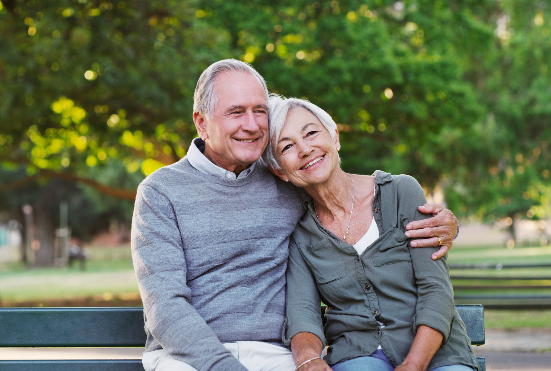 Couple sitting close on a park bench, smiling, arms around each other, with a green background.