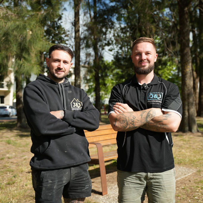 Two men, arms crossed, standing outdoors near a park bench. One in black hoodie, the other in a black polo shirt.