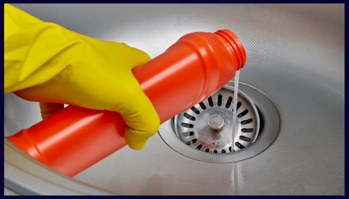 Person wearing yellow gloves pouring chemical drain cleaner into a stainless steel sink in a Canberra home