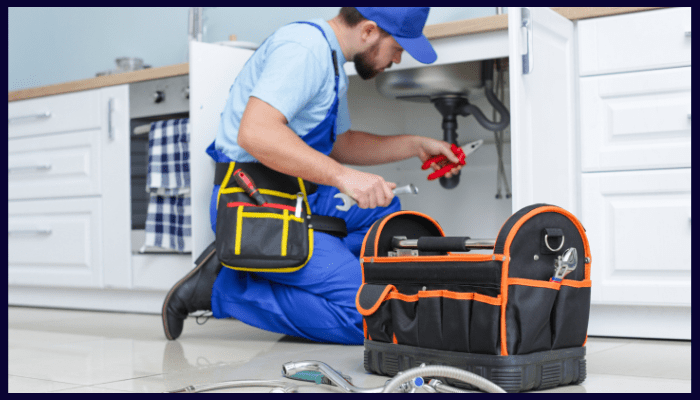 Plumber kneels under kitchen sink, working on pipes with tools. Wearing blue uniform and cap.