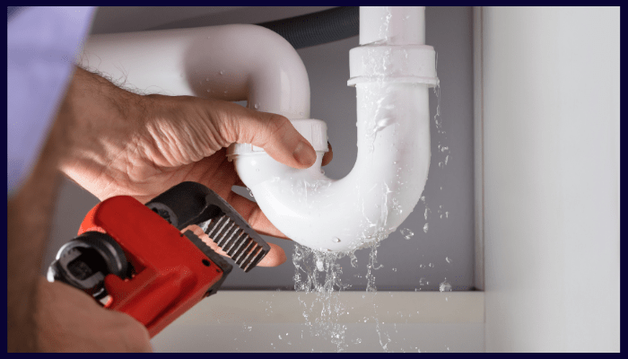 Plumber tightening a wrench on a leaky white pipe under a sink; water is spraying.