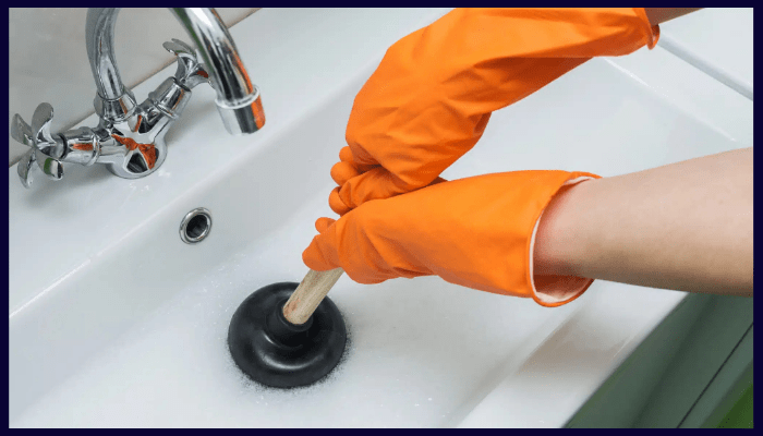 Person wearing orange gloves using a plunger to clear a clogged bathroom sink in a Canberra home