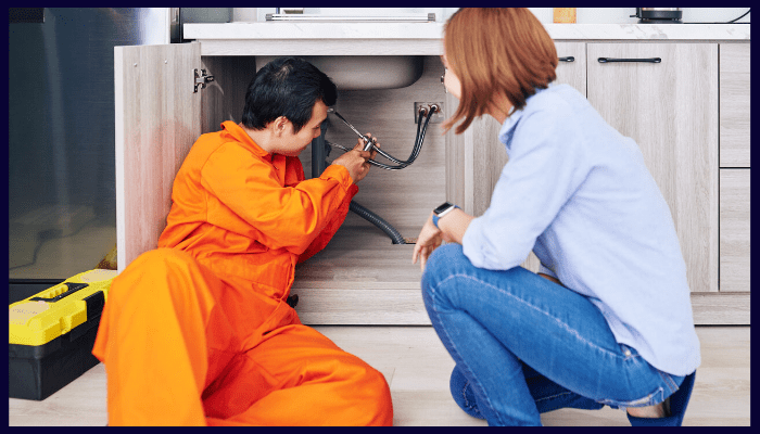 Plumber in orange jumpsuit fixing a sink while a woman watches, in a kitchen.