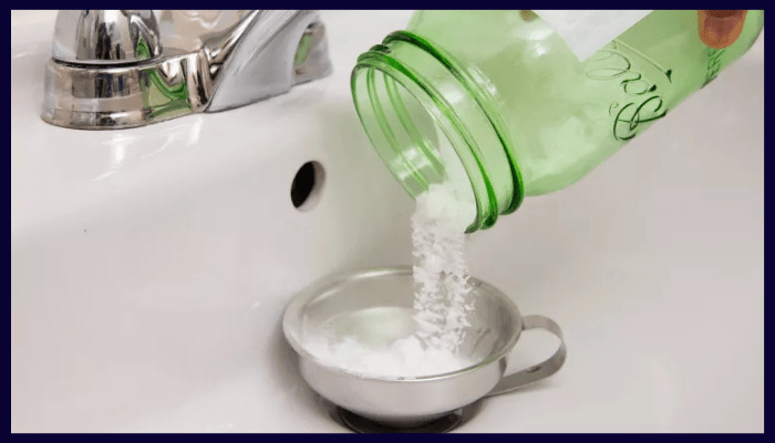 Baking soda being poured from a green jar into a sink drain to illustrate the myth of using baking soda and vinegar for blocked drains.