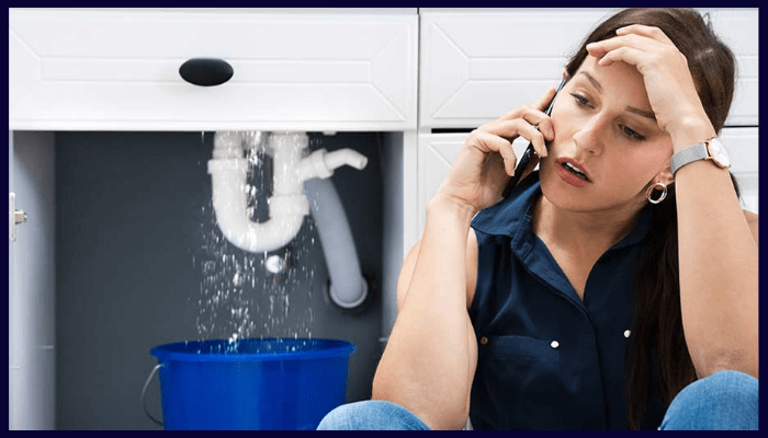Worried woman calling a plumber as water leaks from a pipe under the sink into a blue bucket.