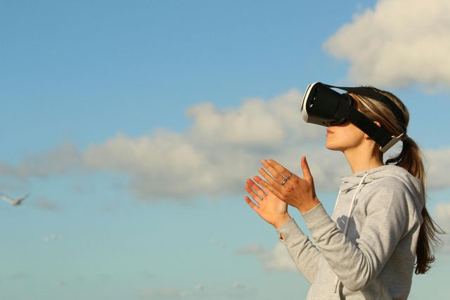 Woman wearing VR headset outdoors with hands raised towards a blue sky and clouds.