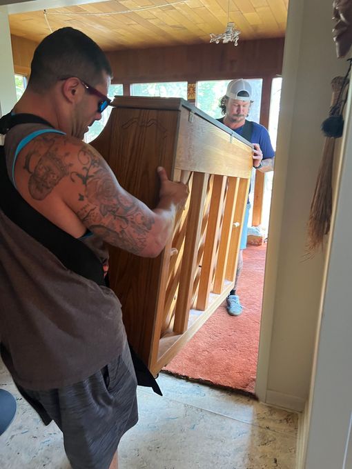 Two people carrying a wooden cabinet through a doorway, one with a moving strap.