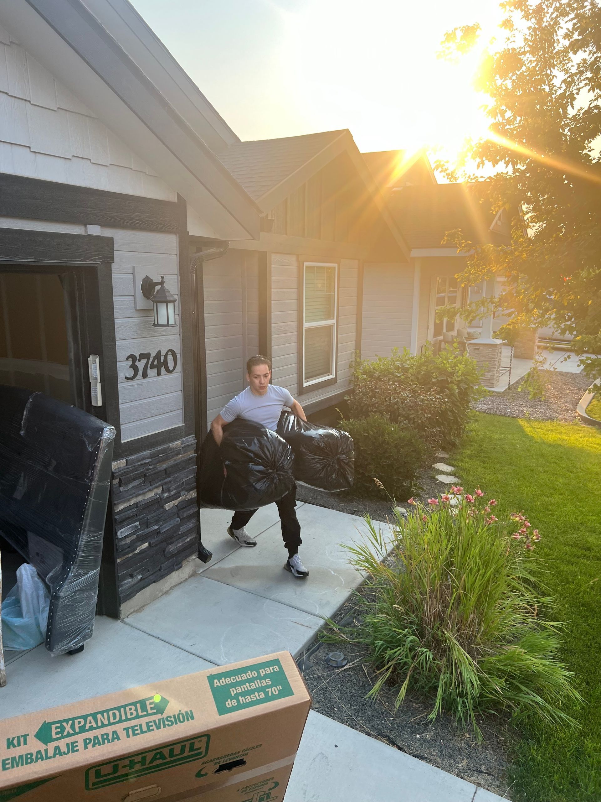 Person carrying black trash bags, standing in front of a house, with a cardboard box and couch visible.