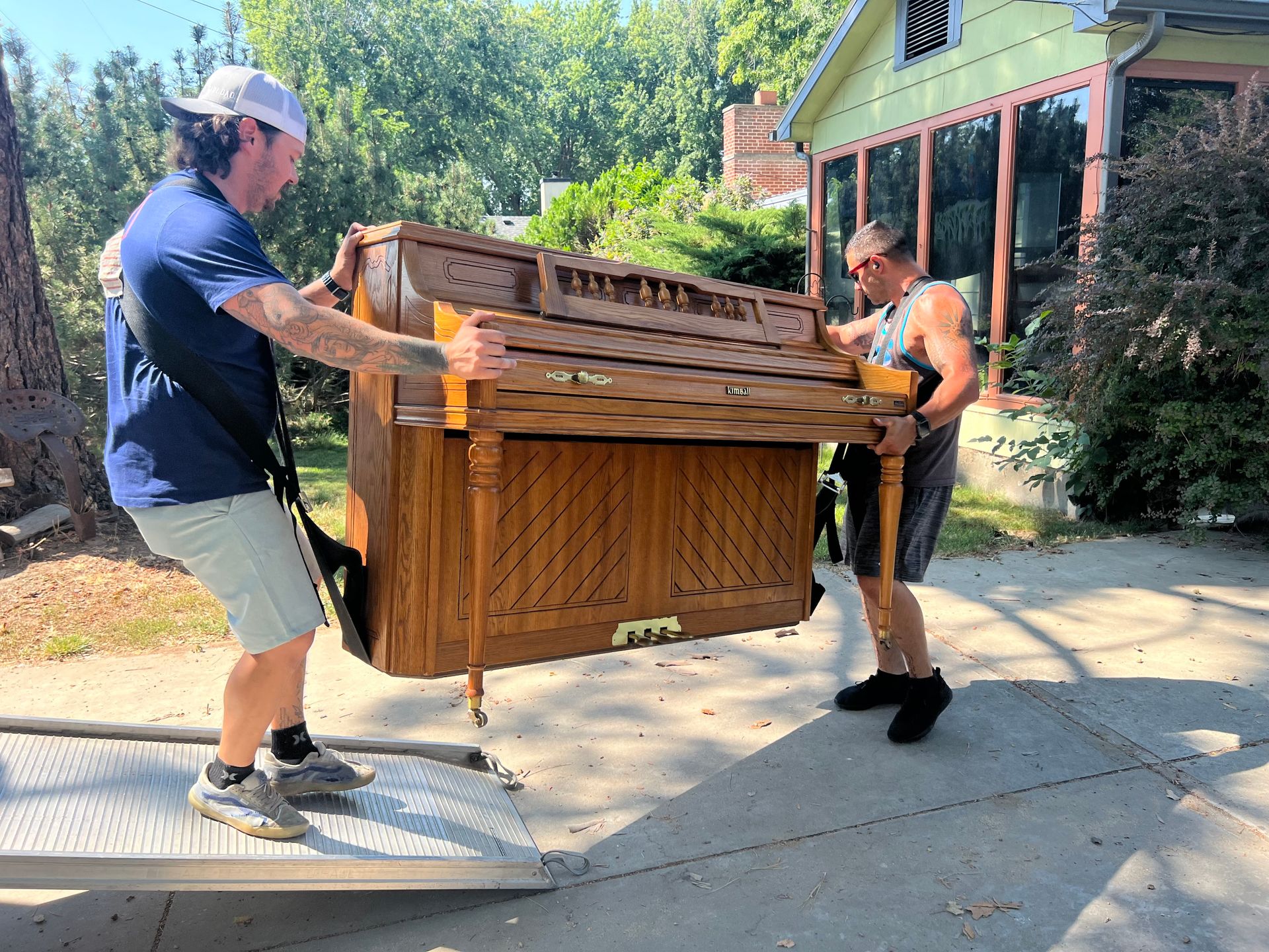 Two men moving a brown upright piano down a ramp. They are outdoors near a house on a sunny day.