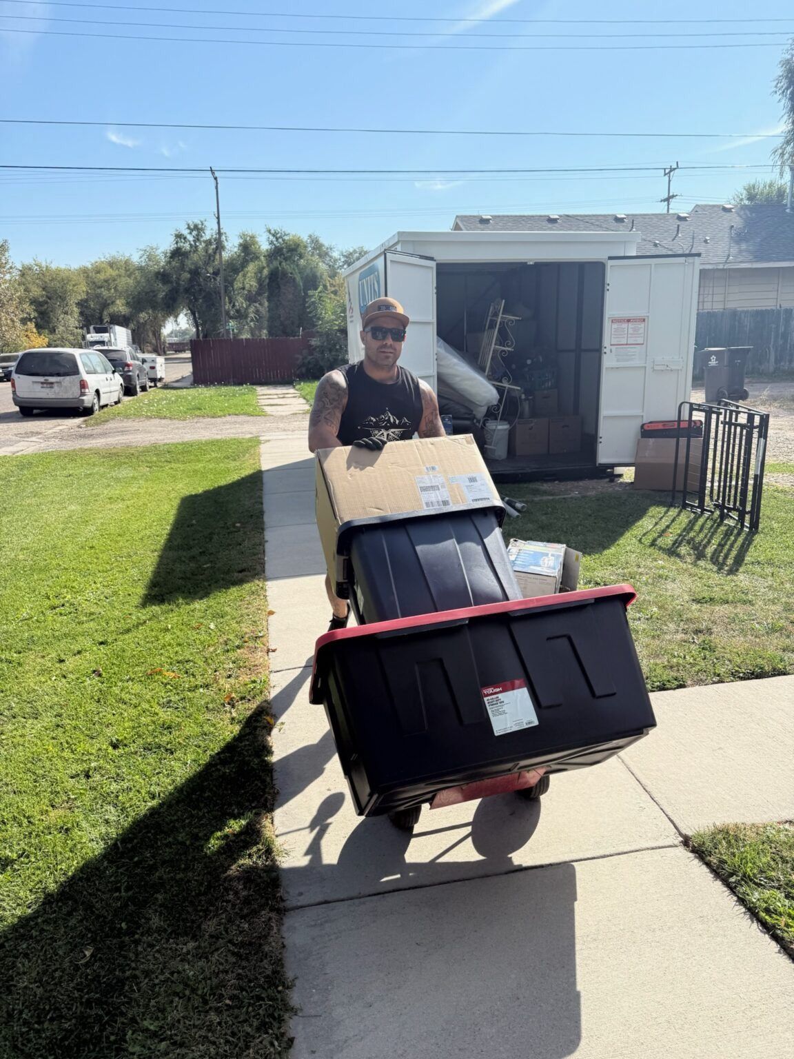 Man pushing a hand truck loaded with moving boxes and bins on a sidewalk. Sunny day.