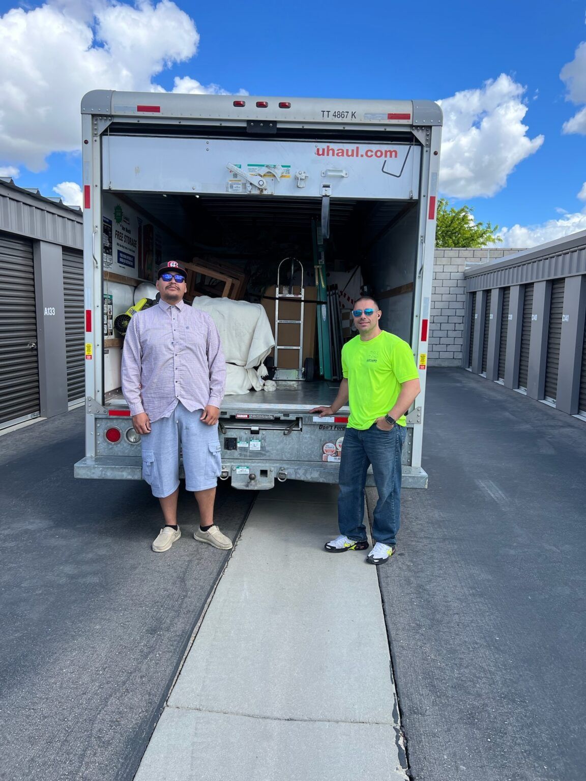 Two men standing in front of a U-Haul truck at a storage facility, truck door open, blue sky.