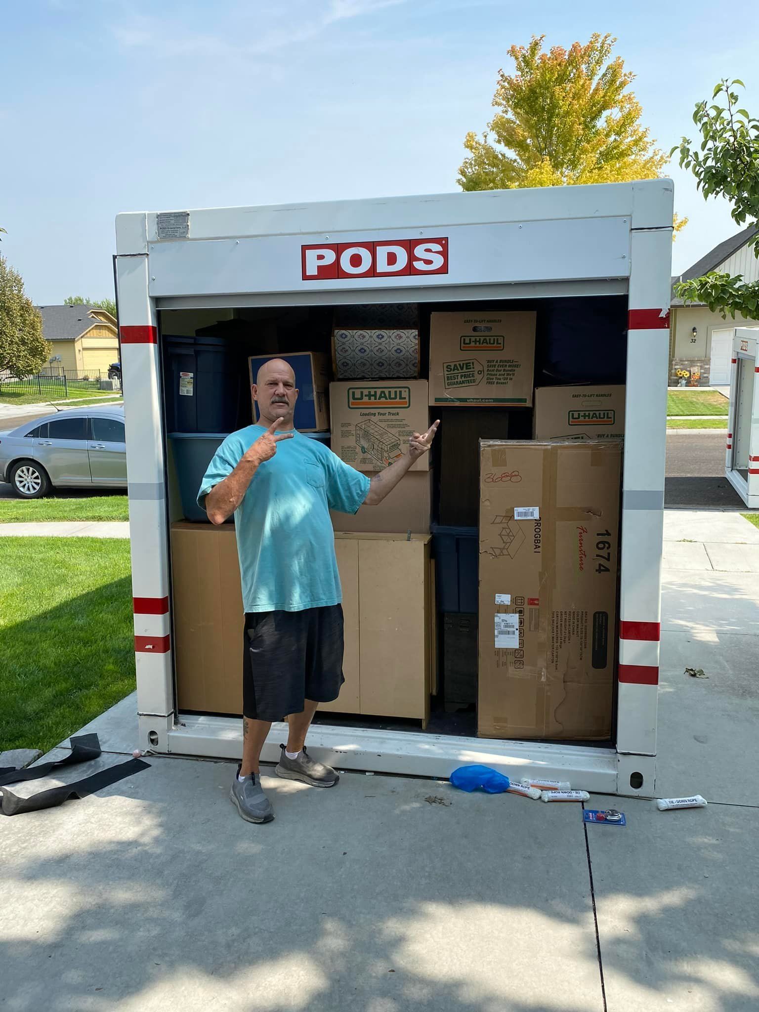 Man stands in front of a PODS container, surrounded by packed boxes. He gestures with his hand, in a residential driveway.