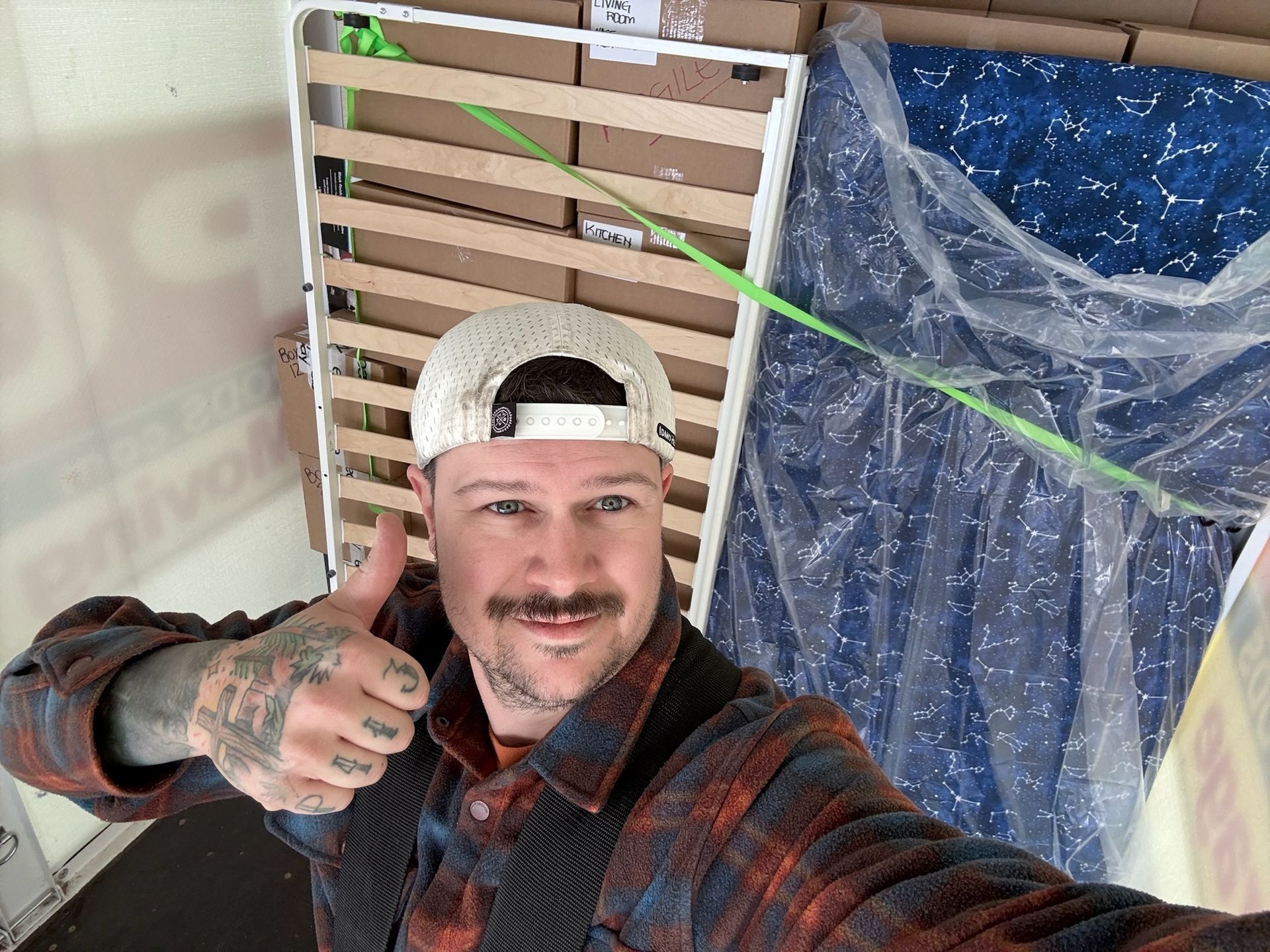 Man in flannel shirt gives thumbs up, surrounded by bed parts in a vehicle.