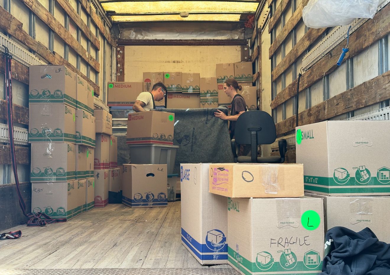 Two people loading boxes into a moving truck. Beige boxes stacked, some with green accents and labels.