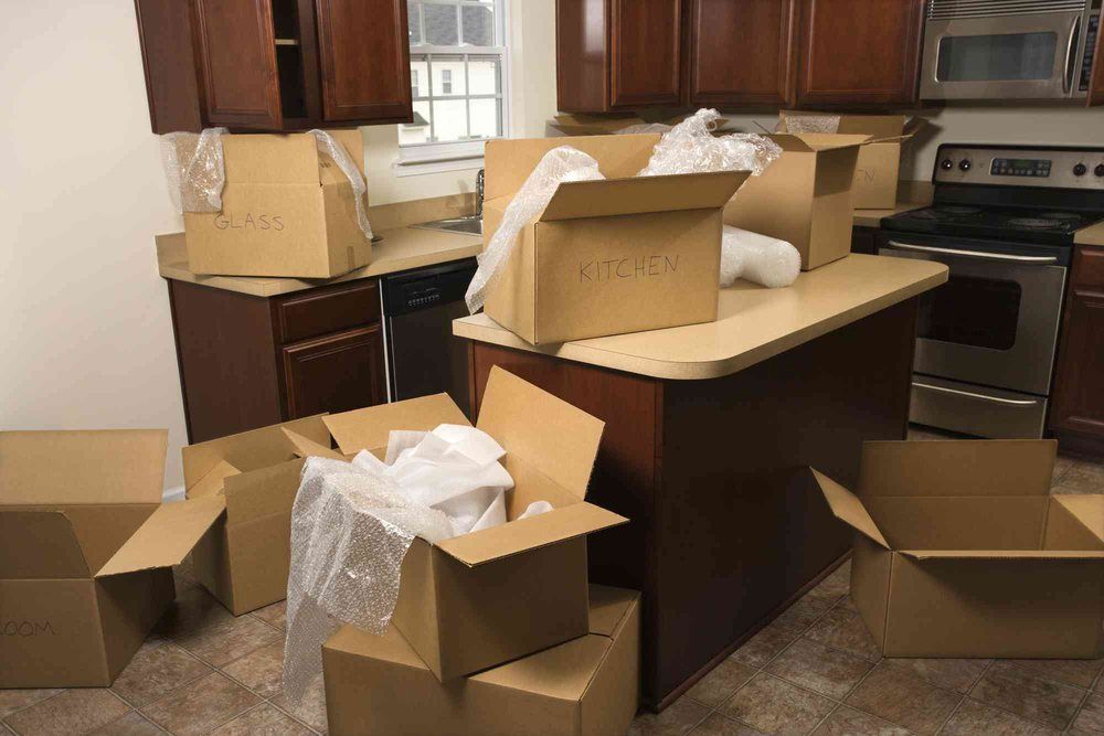 Cardboard boxes in a kitchen, likely for moving. Boxes are on countertops and floor. Brown cabinets and a stove are visible.