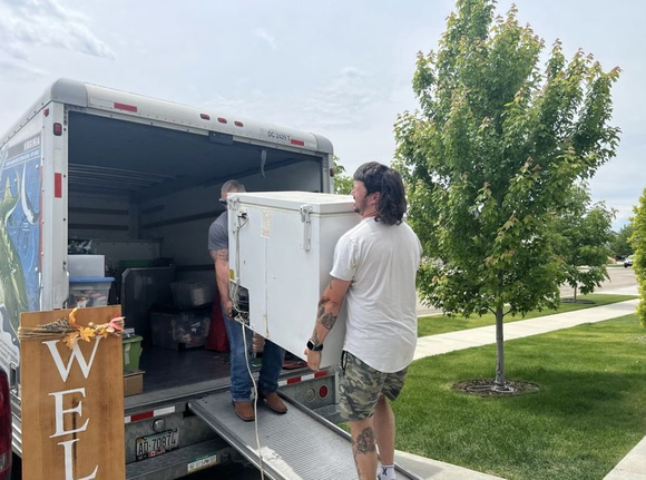 Two people loading a white box onto a moving truck ramp. Outdoors on a sunny day.