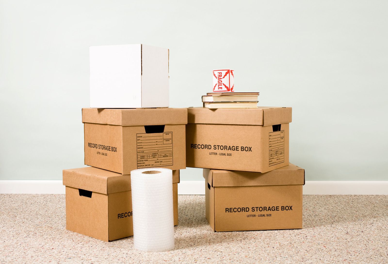 Cardboard boxes stacked for moving, books, hourglass, and bubble wrap roll on carpet.