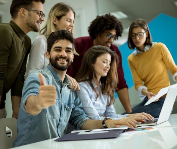 Six people huddled around a laptop, one of them is looking at the camera and gesturing a thumbs-up