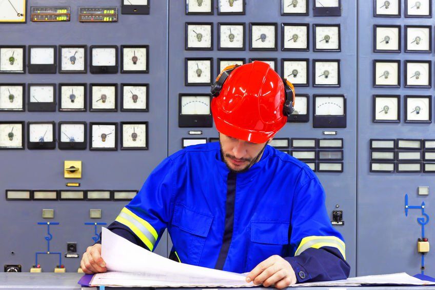 A man wearing a hard hat looking at a draft sheet. Behind him is a machine with many pressure gauges.