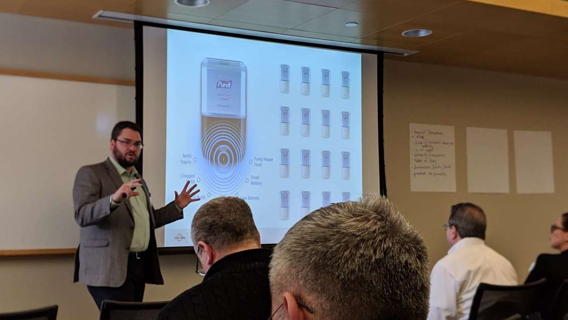 A man giving a presentation. On the projector screen is a motion activated hand sanitizer machine.