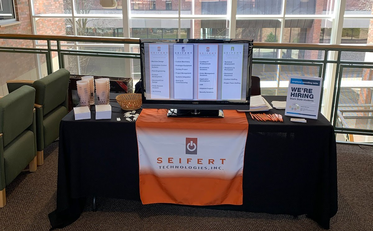 Seifert Technologies info table, with cups, business cards, pencils, monitor, and a display card with 