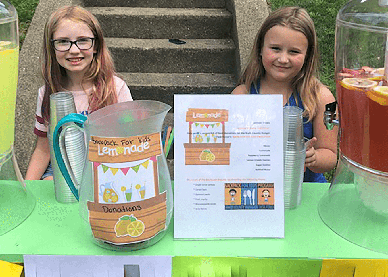 Two children attending a lemonade stand. There is a pitcher filled with donations and two flavors of lemonade.