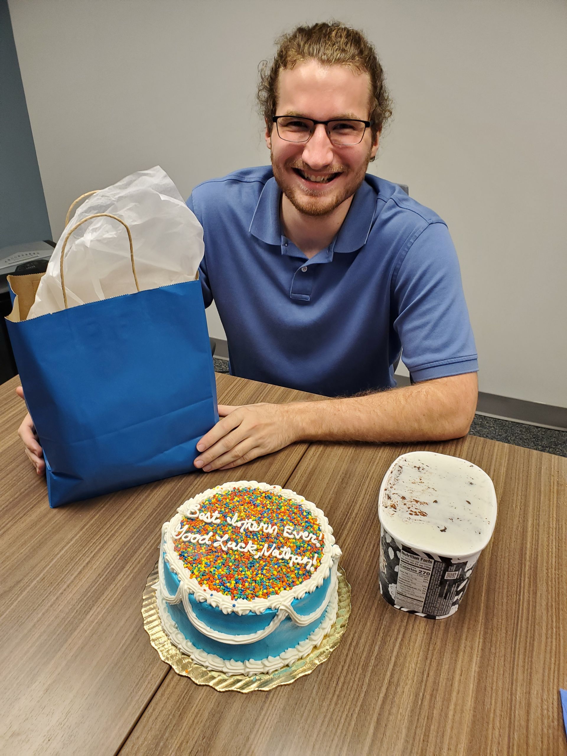 Nathan smiling, holding a gift bag, at a table. In front of him is a cake and ice cream.