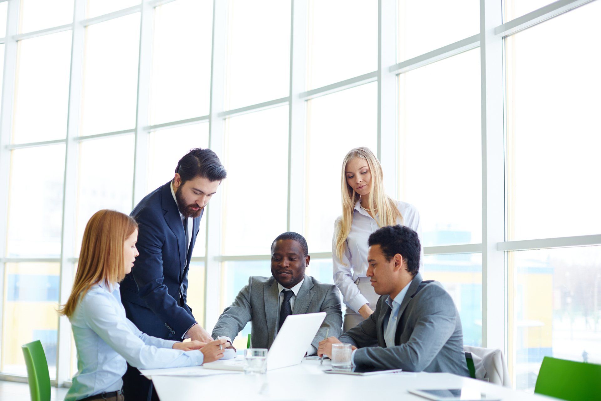 Five people in business wear at a table, deliberating around a laptop.