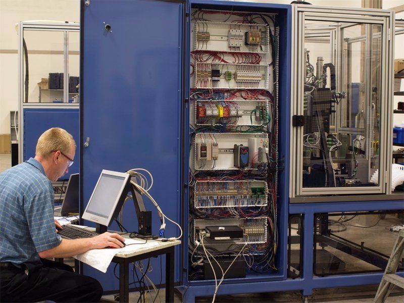 Man at a computer, next to a PLC panel in a warehouse.