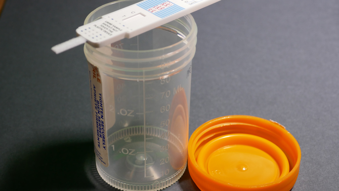 A drug test cup with an orange lid and an integrated test panel resting on top of the container.