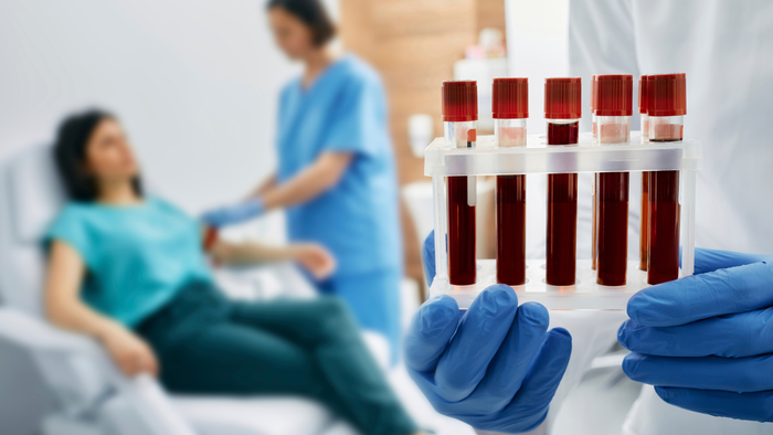 A person in blue gloves holds a rack of blood collection tubes, with a medical professional attending to a patient nearby.