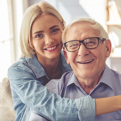 A young person in a denim shirt smiles and hugs an older person wearing glasses and a light purple button-down shirt.