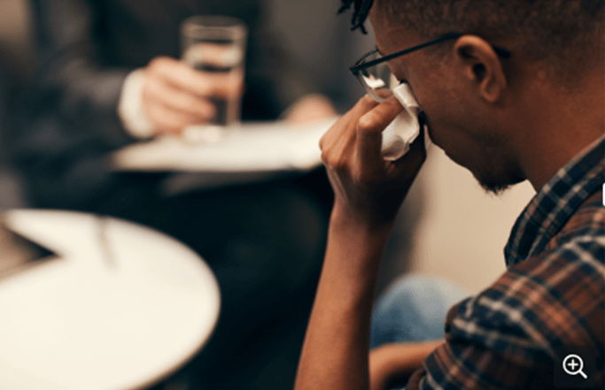 A person wearing glasses holds a tissue to their eyes, sitting opposite someone holding a glass of water during a session.