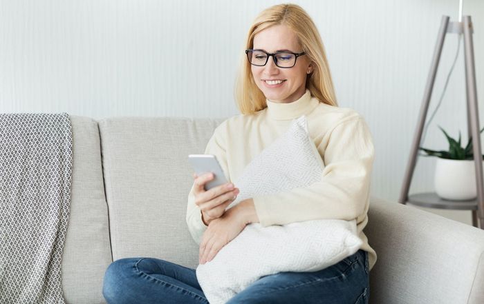 A smiling person with glasses wearing a beige sweater sits on a gray couch, holding a smartphone and a white pillow.