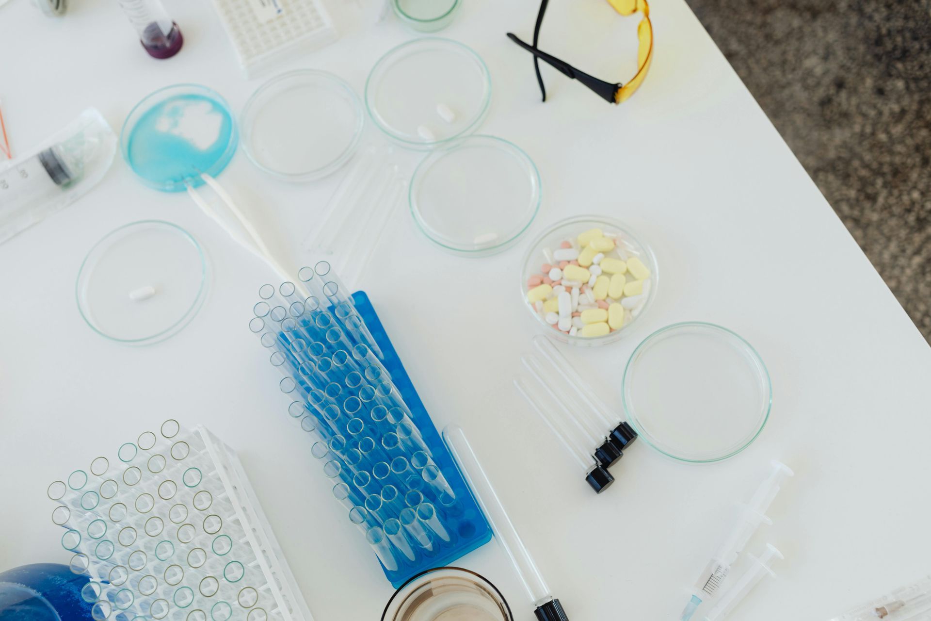A white lab table holds petri dishes, a blue pipette rack, test tubes, safety goggles, and assorted colorful pills.