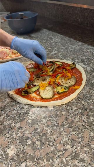 Mani guantate aggiungono i condimenti a una pizza cruda su un piano di lavoro in granito.