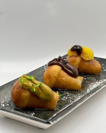Tre pasticcini di piccole dimensioni, con guarnizione di cioccolato e verde, disposti su un piatto rettangolare nero.
