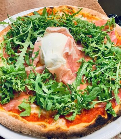 Pizza condita con rucola, prosciutto crudo e una cucchiaiata di burrata su un piatto bianco.