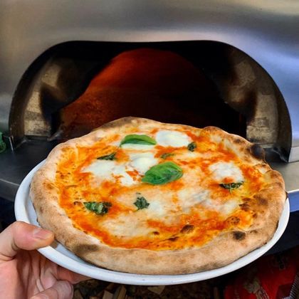 Una mano regge un piatto bianco con una pizza margherita all'interno di un forno a legna.