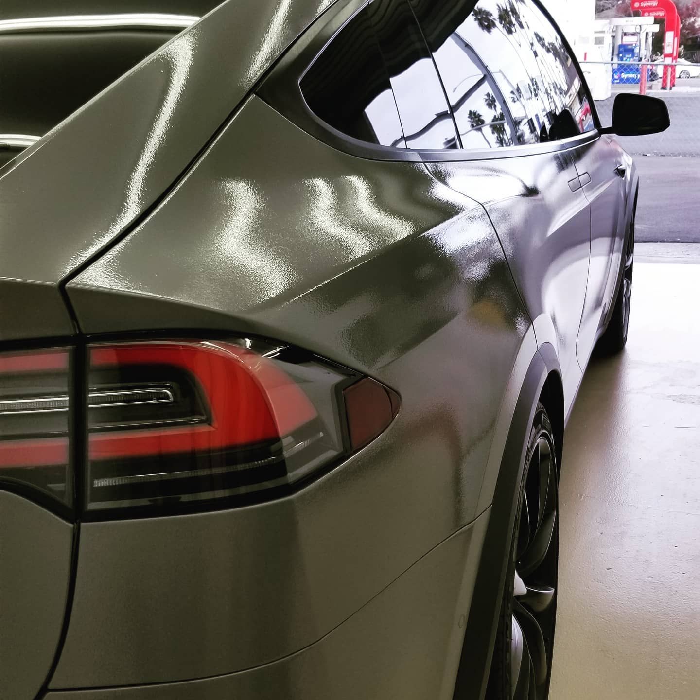Gray Tesla SUV, close-up rear view, showing taillight, window, and wheel. Shiny paint, outdoors.