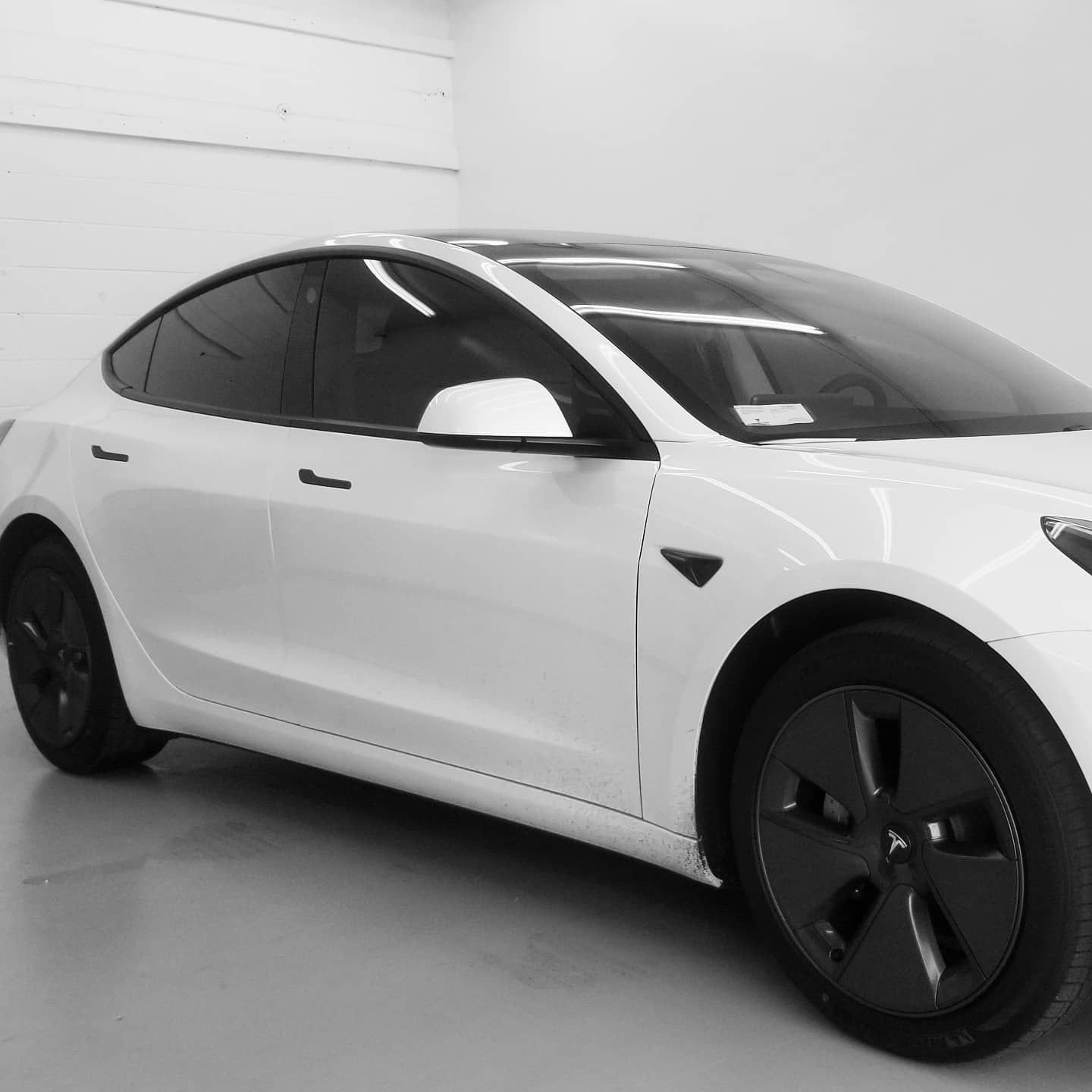 White Tesla Model 3 electric vehicle parked in a white-walled garage with tinted windows.