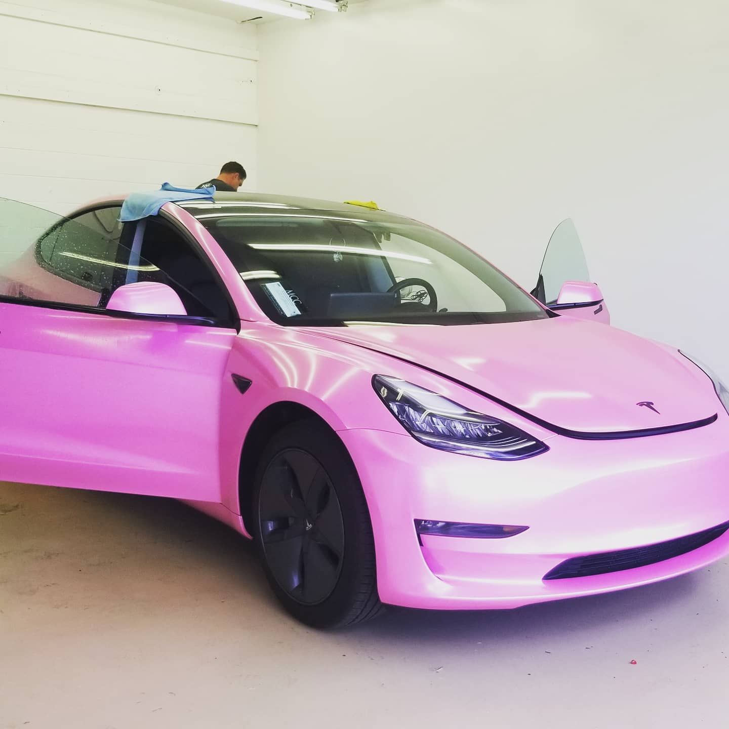 A pink Tesla car being worked on by a person in a white room, with the door and hood open.