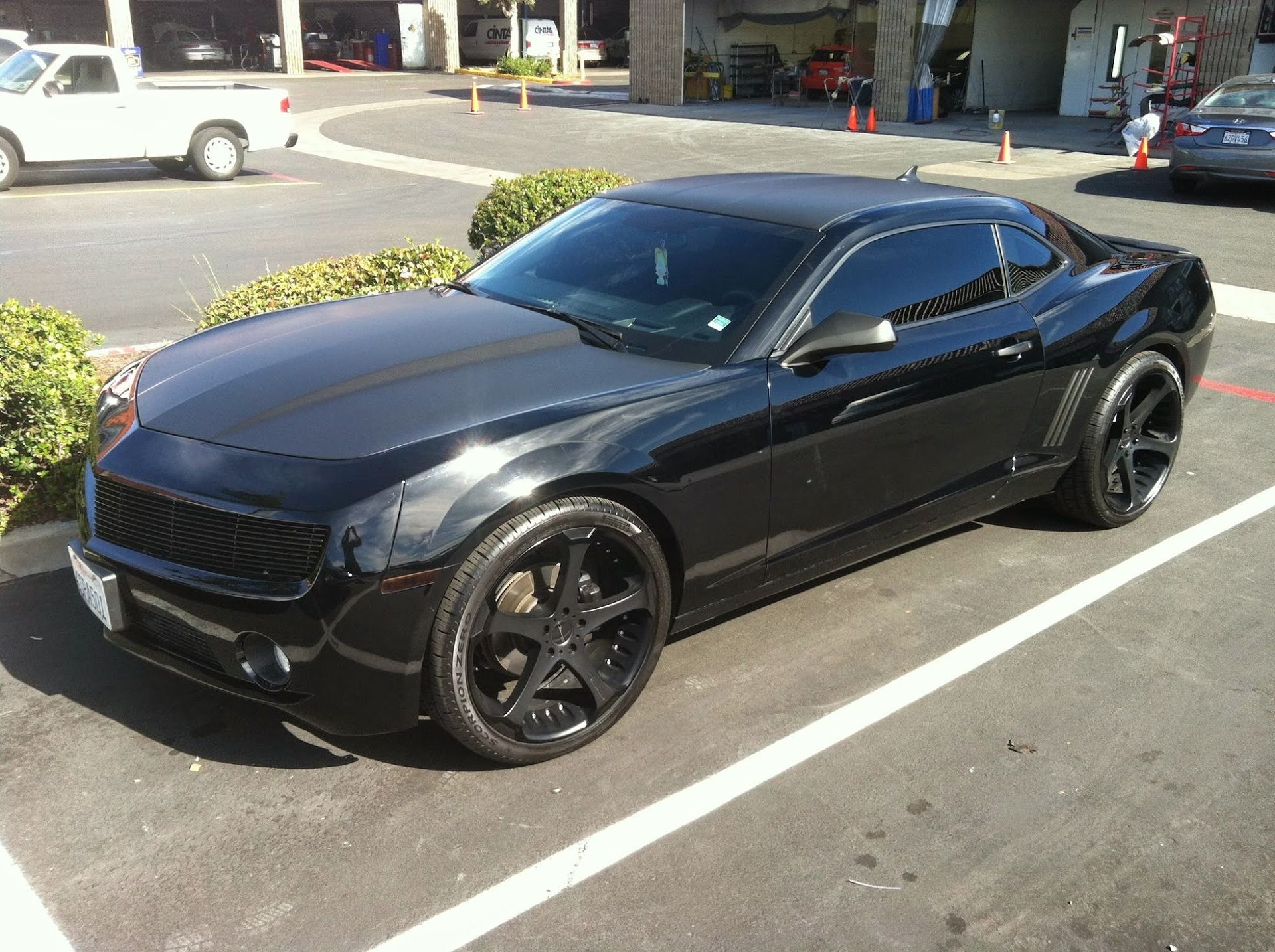Black modified sports car parked in a sunny lot. Black wheels, tinted windows, and hood cover.
