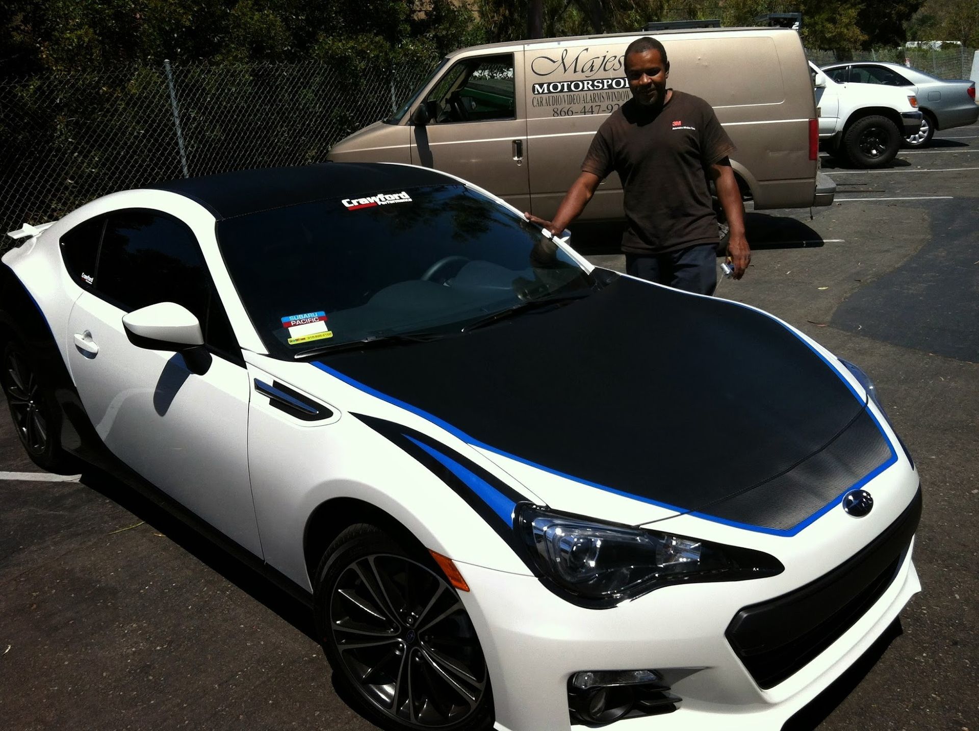 Man standing beside a white Subaru BRZ with a black hood and blue stripes, parked in a lot.