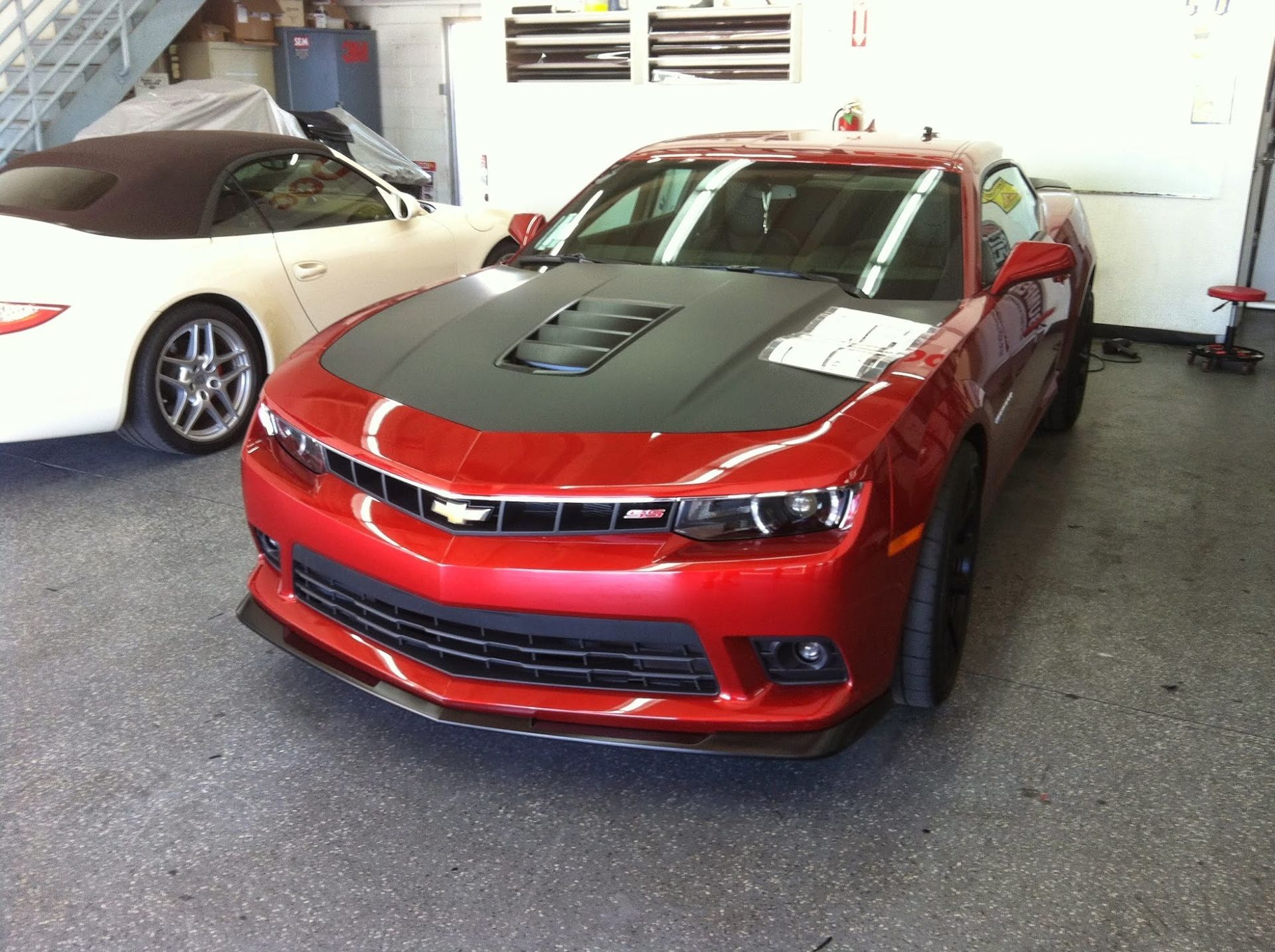 Red Chevy Camaro with black hood in a garage, next to a white convertible.
