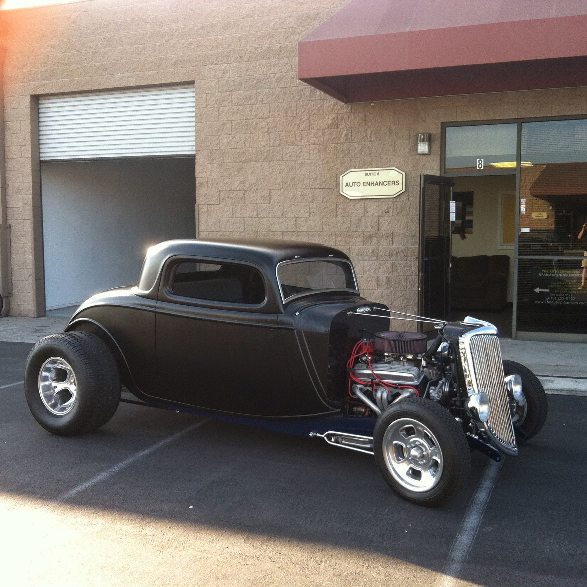 Black hot rod car parked outside a building with an open garage door.
