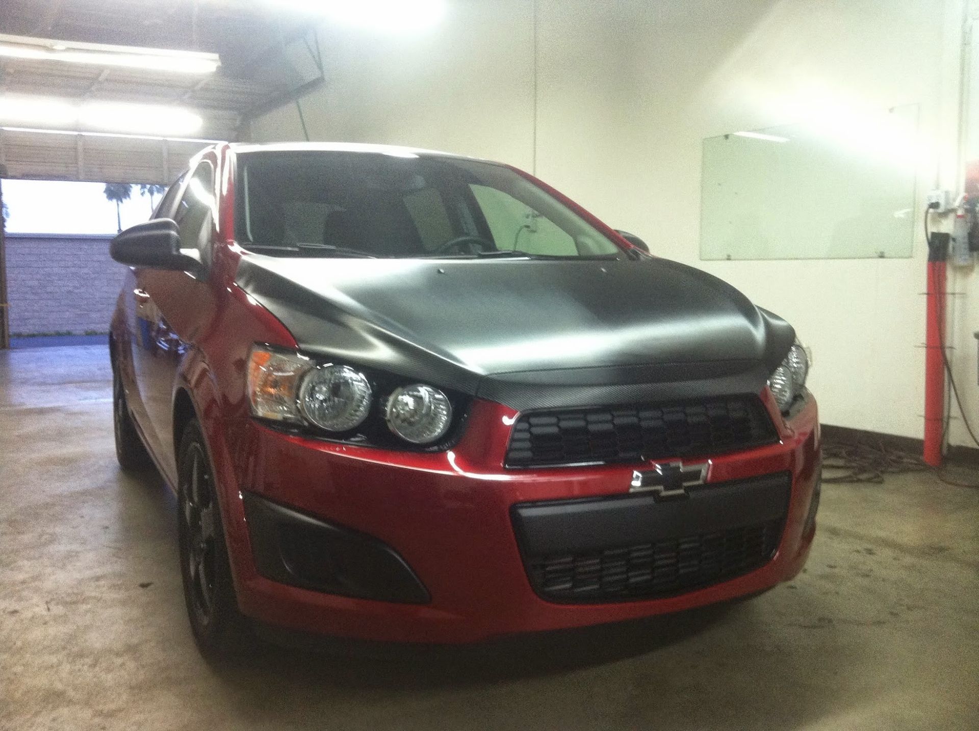 Red Chevy Sonic with a black hood parked in a garage.