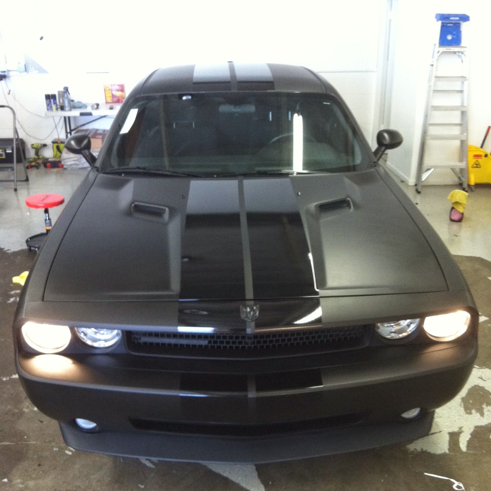 Black Dodge Challenger with racing stripes in a garage, headlights on.