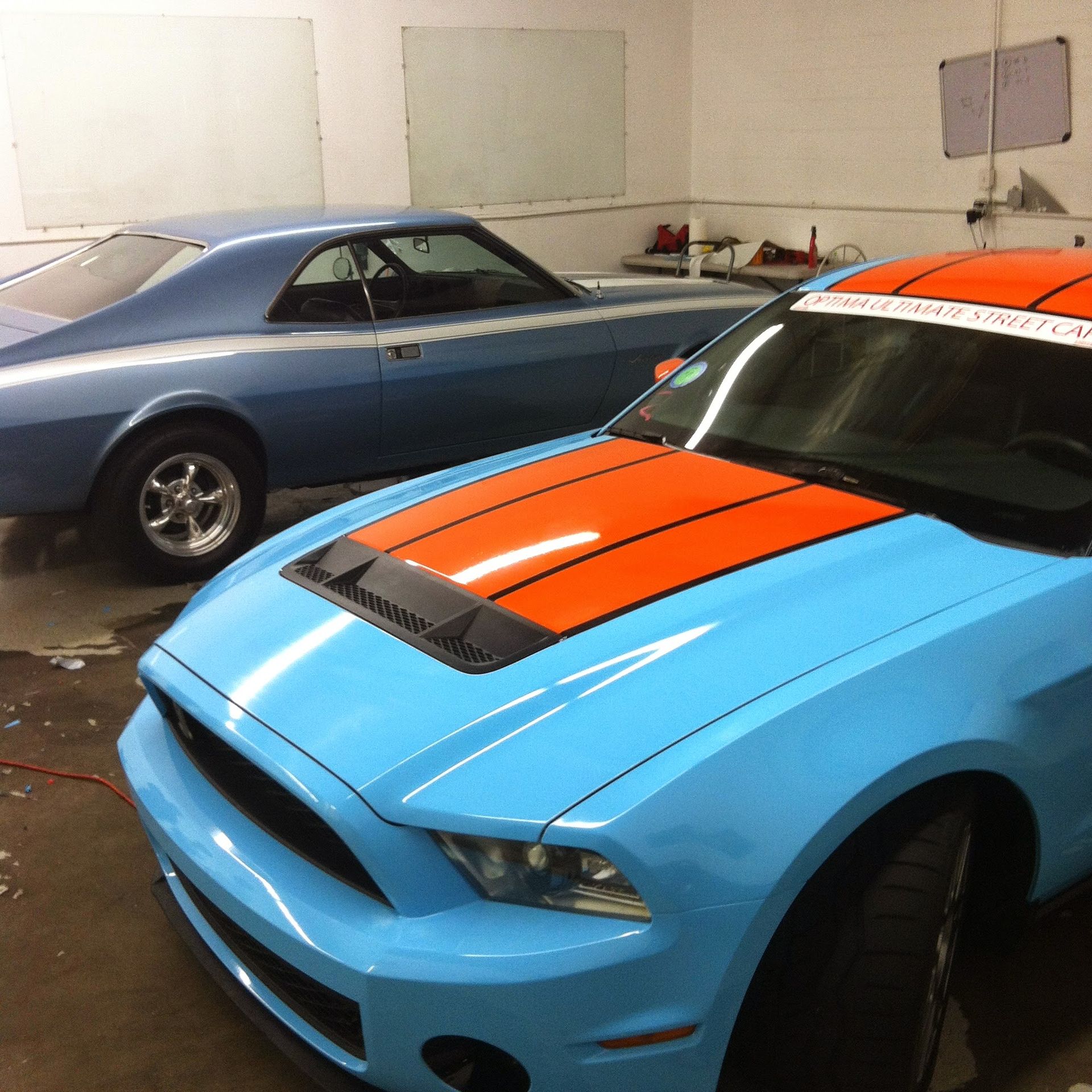 Two blue cars, one classic and one modern, in a garage. The modern car has an orange stripe.