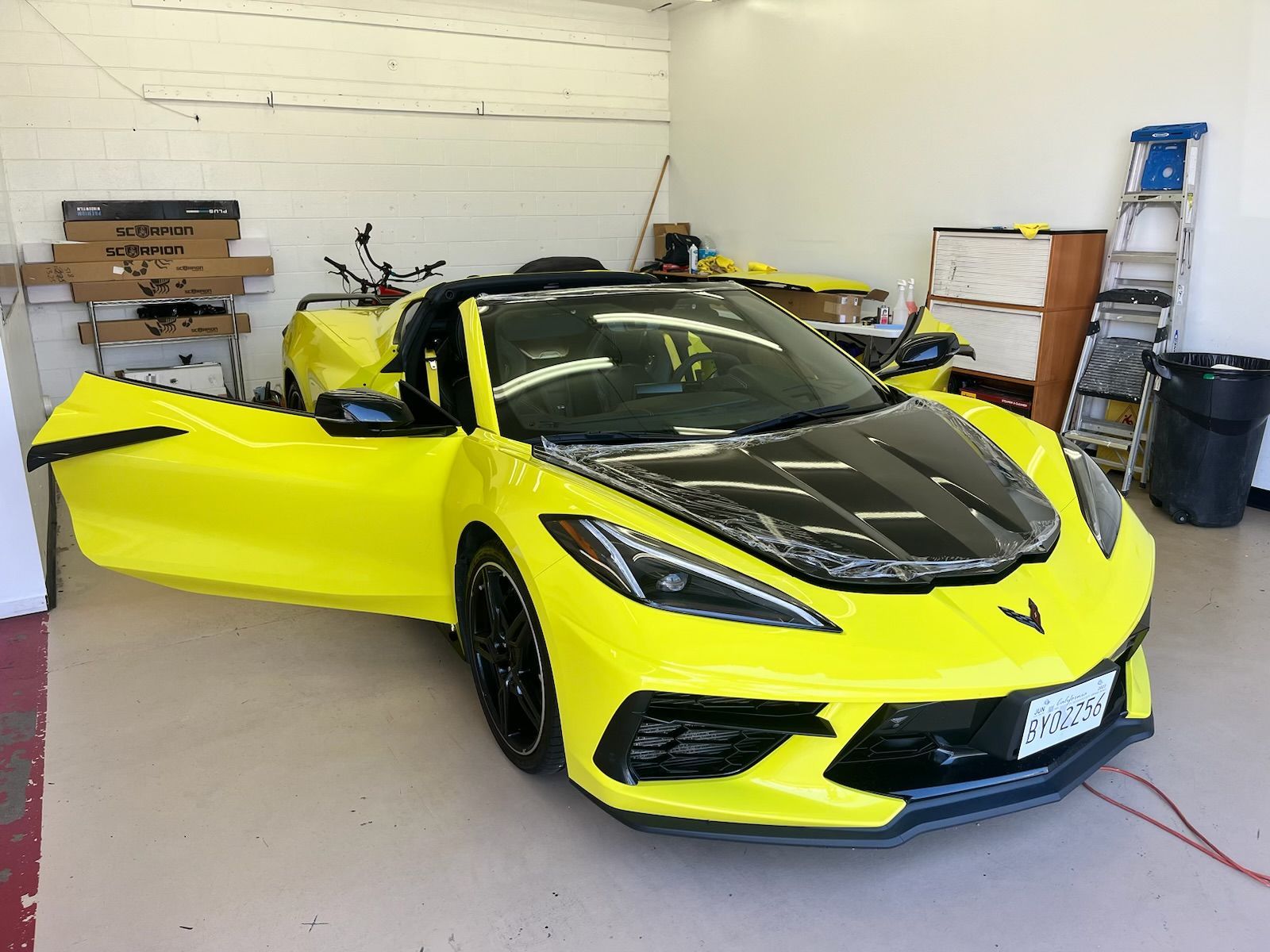 Yellow convertible sports car with the door open, inside a garage. Black hood, black wheels.